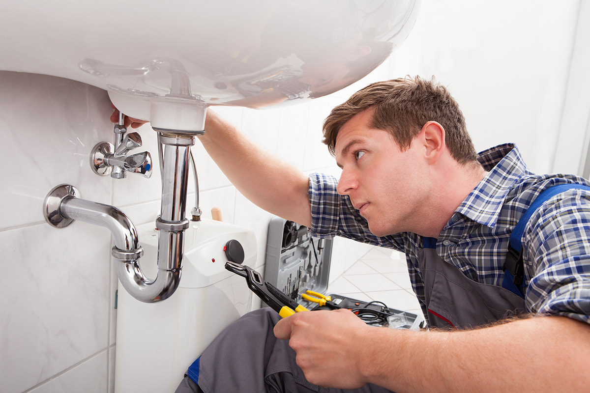 A professional plumber in a plaid shirt and overalls is carefully repairing the pipes under a white bathroom sink using a wrench.